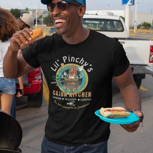 Könnte beinhalten: Schwarzes T-Shirt mit einem "Lil' Pinchy's Cajun Kitchen"-Motiv. Das Design umfasst einen Cartoon-Flusskrebs in einem Topf, Text und den Satz "The best crawfish in Louisiana!". Die Person trägt eine blau-schwarze Trucker-Mütze.