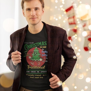 May include: A person wearing a black t-shirt with a Christmas tree graphic and the text "Cletus Woods Christmas Tree Farm". The person is also wearing a maroon blazer and khaki pants. The background has Christmas decorations.