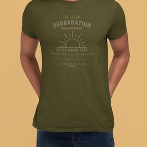 May include: An olive green short-sleeve t-shirt featuring a graphic with the text THE SOLAR DEGRADATION DEPARTMENT, SOLAR CURED TEES, Quality tested in open truck beds, and A BRANCH OF GUILTY KICK APPAREL, centered above a sun icon.