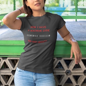 Puede incluir: Camiseta gris oscuro con el texto "NOW I HAVE A MACHINE CODE" en rojo, seguido de código binario y "HO-HO-WHAT?". La camiseta se usa con vaqueros azul claro.