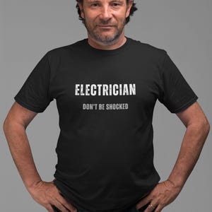 Op de afbeelding: Zwart T-shirt met de witte tekst "ELECTRICIAN DON'T BE SHOCKED". Het shirt heeft korte mouwen en wordt gedragen door een persoon met een zonnebril op het hoofd. De persoon draagt een blauwe spijkerbroek.
