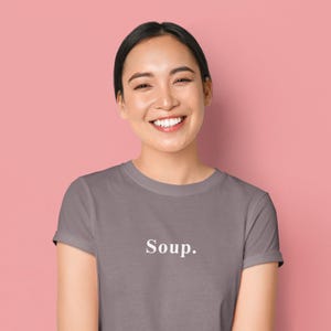 May include: A woman wearing a gray t-shirt with the word "Soup." printed in white. The shirt has short sleeves and a crew neck. The background is a solid pink color.