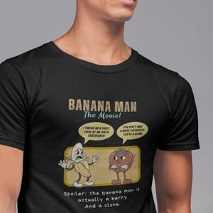 Könnte beinhalten: Schwarzes T-Shirt mit einer Cartoon-Grafik aus "Banana Man: The Movie!". Die Grafik zeigt eine Banane und eine Erdbeere in einer hitzigen Diskussion, mit Sprechblasen und Text, der lautet: "Spoiler: The banana man is actually a berry. And a clone."