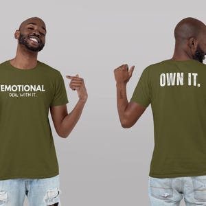 May include: Olive green t-shirt with the text "EMOTIONAL DEAL WITH IT" on the front and "OWN IT" on the back. The t-shirt is worn by a person in blue jeans. The person is pointing at the front of the shirt.