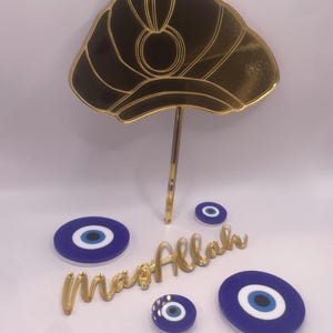 May include: A gold-colored cake topper in the shape of a turban, with a matching stick. Several blue and white evil eye charms and a gold-colored "MashAllah" script are also present. The items are arranged on a white surface.
