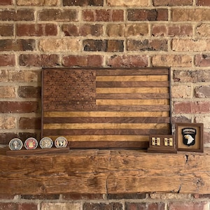May include: A wooden American flag with carved stars and stripes, displayed on a rustic wooden mantel. Several military challenge coins and framed insignia are arranged below the flag, showcasing a patriotic theme.