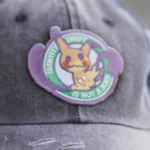 May include: A faded grey baseball cap with a circular patch. The patch features a cartoon character and the text "IDENTITY IS NOT A JOKE". The cartoon character is yellow and orange.
