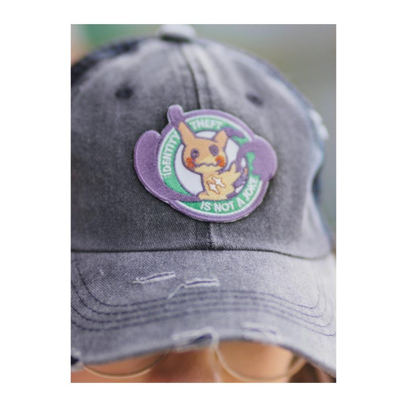 May include: A faded grey baseball cap with a circular patch. The patch features a cartoon character and the text "IDENTITY IS NOT A JOKE". The cartoon character is yellow and orange.