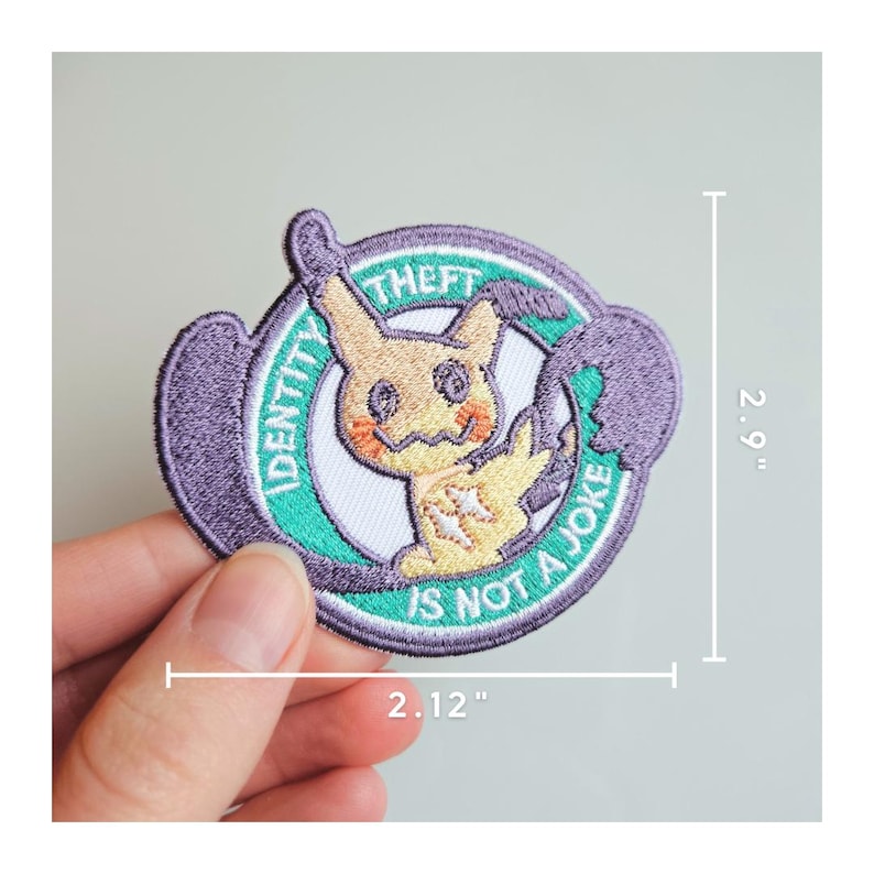 May include: Embroidered patch with a cartoon character and the text "IDENTITY THEFT IS NOT A JOKE." The patch measures approximately 7.37 cm high and 5.38 cm wide. The design incorporates purple, green, yellow, and white.