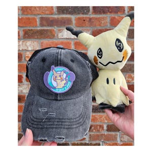 May include: A distressed grey trucker cap with a black mesh back and a circular patch featuring a cartoon character and the text "THEFT IS NOT A JOKE". Next to it is a plush toy of a yellow and black character.