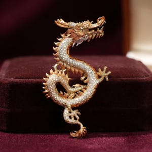 Könnte beinhalten: Goldfarbene Drachenbrosche mit detailliertem Design. Der Drache ist in einer geschwungenen Form dargestellt, sein Körper ist mit kleinen, funkelnden Steinen bedeckt. Kopf und Krallen des Drachen sind aufwendig detailliert.