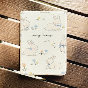 May include: A rectangular, off-white case with a repeating pattern of white bunnies wearing blue bows, flowers, and the text "many bunnies." The bunnies are in different poses, set against a backdrop of grass and flowers.