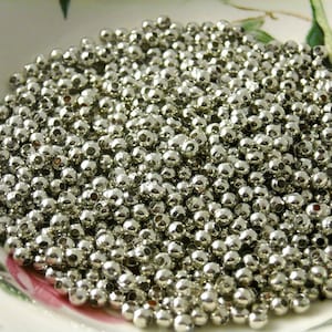 May include: A close-up shot of a pile of small, shiny, silver-colored beads. The beads are round with a small hole through the center. They are scattered in a white and floral patterned bowl. These beads are ideal for jewelry making and crafts.