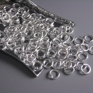 May include: A close-up of a silver tray filled with silver jump rings. The jump rings are scattered on a gray surface.
