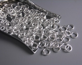 May include: A close-up of a silver tray filled with silver jump rings. The jump rings are scattered on a gray surface.