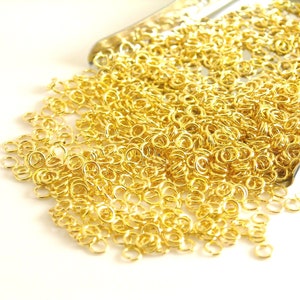 May include: A large quantity of gold jump rings, a type of jewelry finding, are piled in a metal tray. The rings are small and circular, and they are all the same size.
