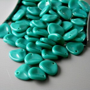 May include: A large pile of teal blue glass beads in the shape of petals. The beads are arranged in a loose pile on a silver surface.