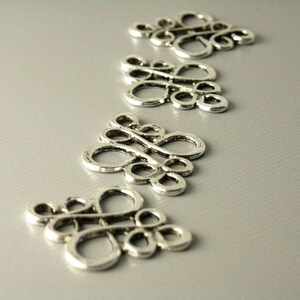 May include: Four silver metal decorative charms in the shape of a stylized knot or swirl design. The charms are arranged in a row on a gray surface.