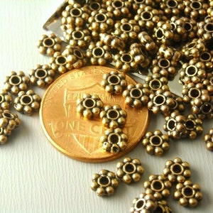May include: Close-up of antique brass-colored flower-shaped beads. The beads are scattered around a penny, showing their size and detail. These small beads are ideal for jewelry making and crafting projects.