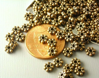May include: Close-up of antique brass-colored flower-shaped beads. The beads are scattered around a penny, showing their size and detail. These small beads are ideal for jewelry making and crafting projects.