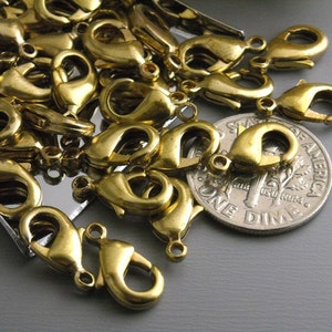 May include: A close-up of a pile of gold-toned metal lobster claw clasps, some of which are laying on top of a US dime coin. The clasps are used for jewelry making.