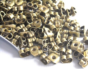 May include: A close-up shot of a pile of small, brass-colored earring backs. The earring backs are in various shapes, including butterfly and barrel styles. The metal has a slightly aged appearance, suggesting a vintage or antique aesthetic. The earring backs are scattered.