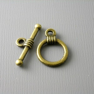 May include: A pair of antique gold-toned metal toggle clasps for jewelry making. One clasp is a bar with a ring on one end and a round knob on the other. The other clasp is a ring with a decorative design.