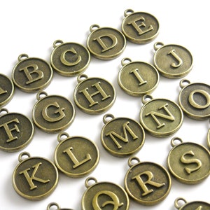 May include: A collection of round, antique bronze-toned metal charms, each featuring a raised capital letter of the alphabet from A to Y. Every circular charm includes a small loop at the top for attachment, displaying a metallic finish.