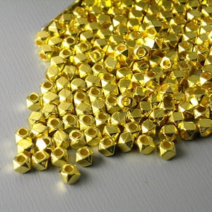 May include: A collection of small, shiny, gold-colored beads. The beads have a geometric, faceted shape and are scattered across a surface. The beads appear to be made of metal and reflect light, creating a bright, eye-catching display.