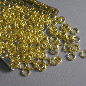 May include: A large quantity of gold jump rings in a metal tray. The jump rings are scattered on a gray surface.