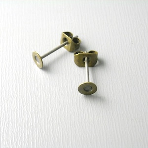 May include: Two gold-toned earring posts with a silver post and a round back. The earring posts are on a white textured surface.