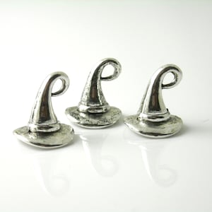 May include: Three silver-colored witch hat charms. Each hat has a curved, pointed top and a wide brim. The charms are made of a metallic material and are displayed on a white surface. These charms are suitable for jewelry making or craft projects.