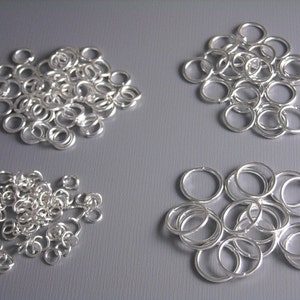 May include: Four piles of silver jump rings of different sizes. The jump rings are arranged in a grid pattern on a grey surface.