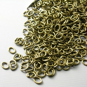 May include: A large pile of gold-toned jump rings, a type of metal ring used in jewelry making. The rings are oval-shaped and are scattered on a white surface.