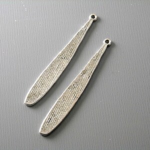 May include: Two silver-toned metal earring blanks with a textured surface. The blanks are long and narrow, with a loop at the top for attaching earring findings.