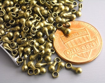 May include: A pile of antique gold-toned metal teardrop-shaped beads with a loop at the top. The beads are scattered on a gray surface next to a US penny.