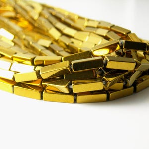 May include: Close-up of a gold-colored necklace. The necklace is made of rectangular, metallic links. The links are arranged in a curved pattern, with the ends of the necklace out of frame. The background is white.