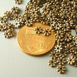 May include: A close-up of a pile of gold-toned metal beads. The beads are small and round, with a raised design that resembles a flower. The beads are scattered on a light-colored surface, and a US penny is visible in the background.