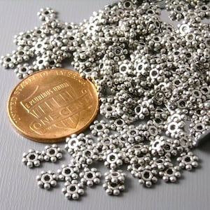 May include: A collection of small, silver-colored metal flower-shaped beads. A U.S. one-cent coin is included for size comparison. The beads have a detailed, textured surface and are scattered across a light gray surface.