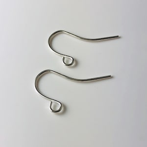 May include: Two silver-colored earring hooks are displayed against a white background. Each hook has a curved design with a small loop at the bottom and a straight, pointed end. The hooks are made of a shiny, metallic material.