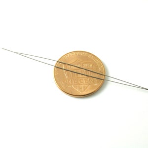 May include: Two thin, silver-colored needles laid out on a white surface, with a US penny placed between them. The penny is facing heads up, with the words "United States of America" and "One Cent" visible.