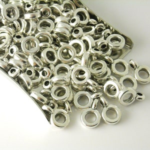 May include: A large pile of silver jump rings with a small loop on one side. The rings are scattered on a white surface.