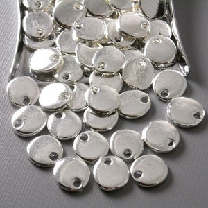 May include: A pile of silver-toned metal round charms with a hole in the center. The charms are scattered on a gray surface.