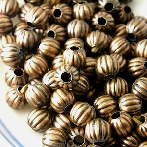 May include: A close-up of a pile of round, copper-colored beads with a ribbed texture. The beads have a hole in the center for stringing.