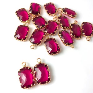 May include: A group of 18 gold-toned, faceted, rectangular, pink glass charms with a loop on each end. The charms are scattered on a white surface.