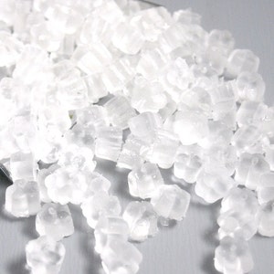 May include: A close-up of a pile of clear, translucent, sugar crystals. The crystals are small and uniform in size, and they are scattered on a light gray surface.