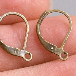 May include: Two gold-toned earring hooks, also known as ear wires, are shown on a hand. The hooks are made of metal and have a smooth, polished finish.