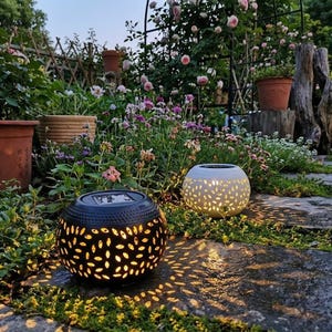 May include: Two solar-powered garden lanterns with a spherical design. One is black, the other is white. Both have a perforated pattern that casts light onto the stone pathway. The lanterns are surrounded by greenery and flowers.