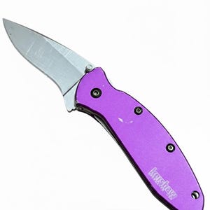 May include: A purple folding knife with a silver blade. The knife has a purple handle with three black rivets and the brand name "Kershaw" printed on the handle.