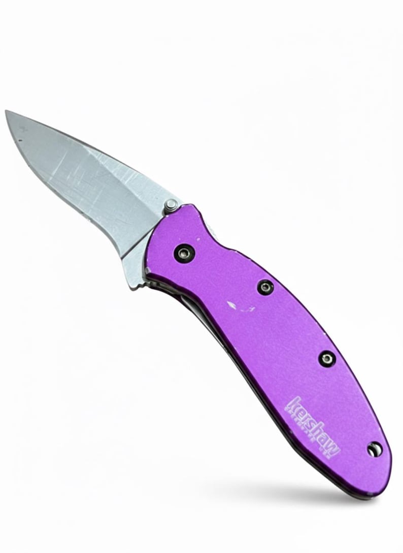 May include: A purple folding knife with a silver blade. The knife has a purple handle with three black rivets and the brand name "Kershaw" printed on the handle.